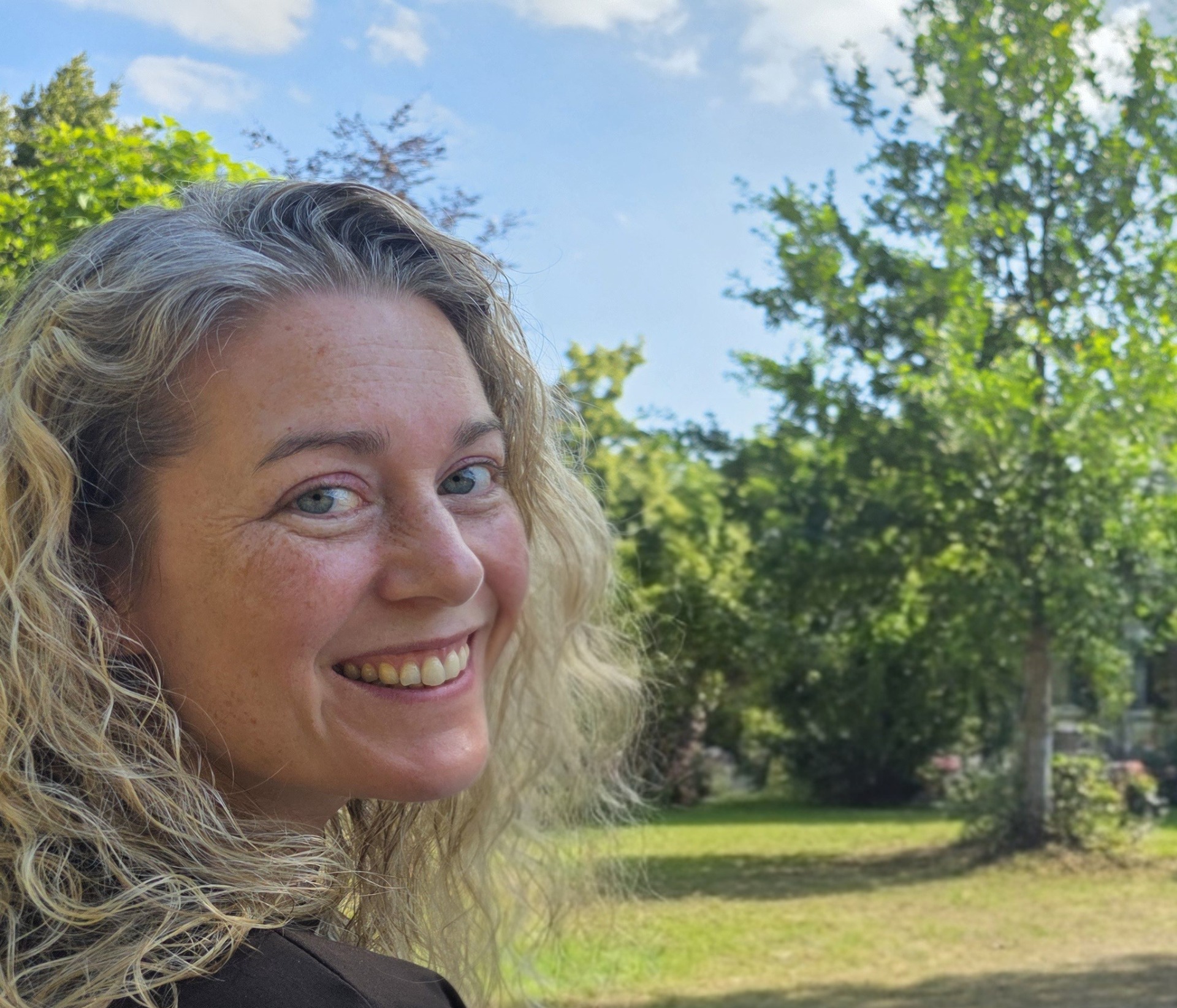Glimlachend gezicht van een vrouw in close-up, buiten in een park met groene bomen en blauwe lucht.